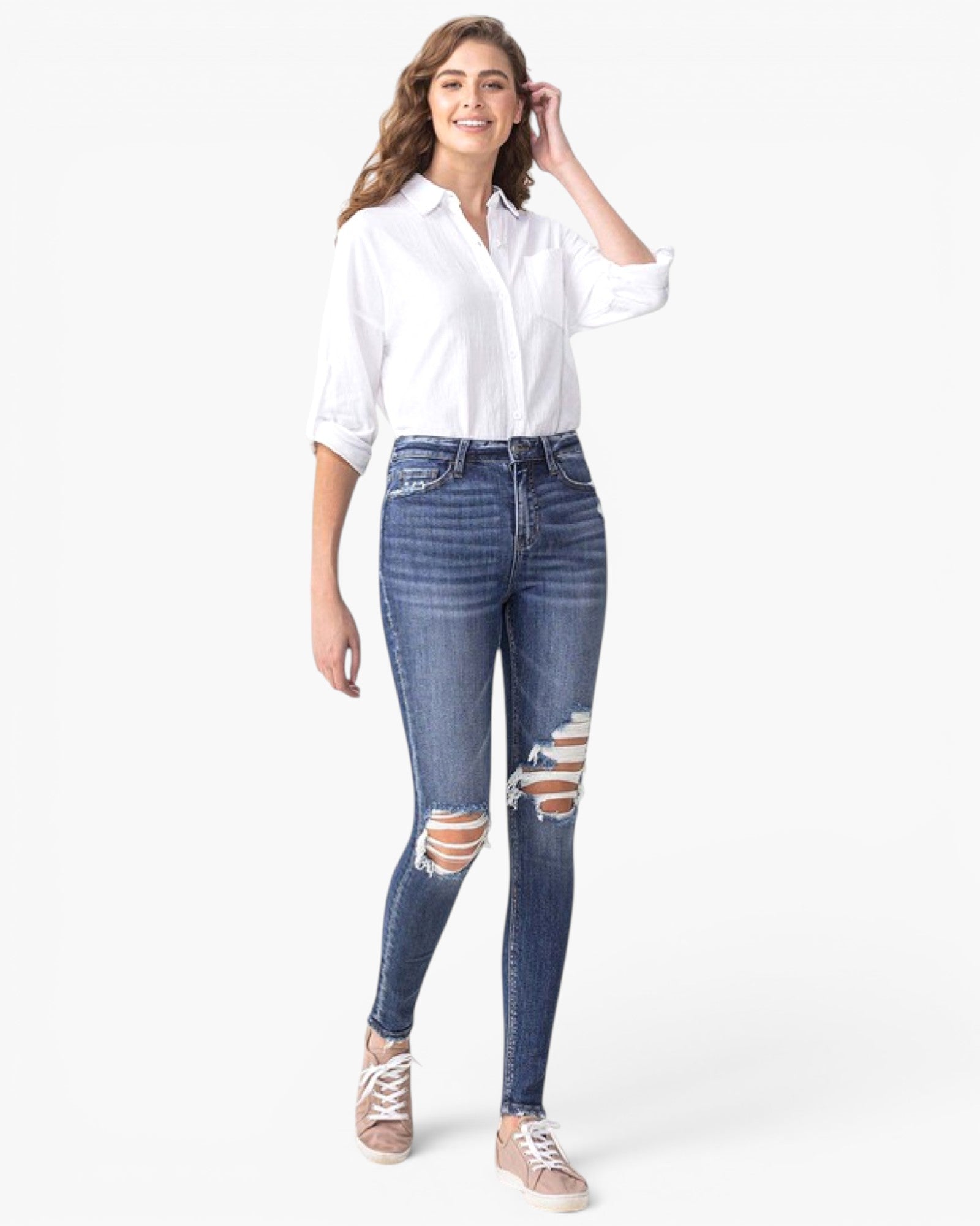 A woman modeling a white button-down shirt and blue distressed skinny jeans with ripped knees, paired with beige sneakers, against a plain white background.