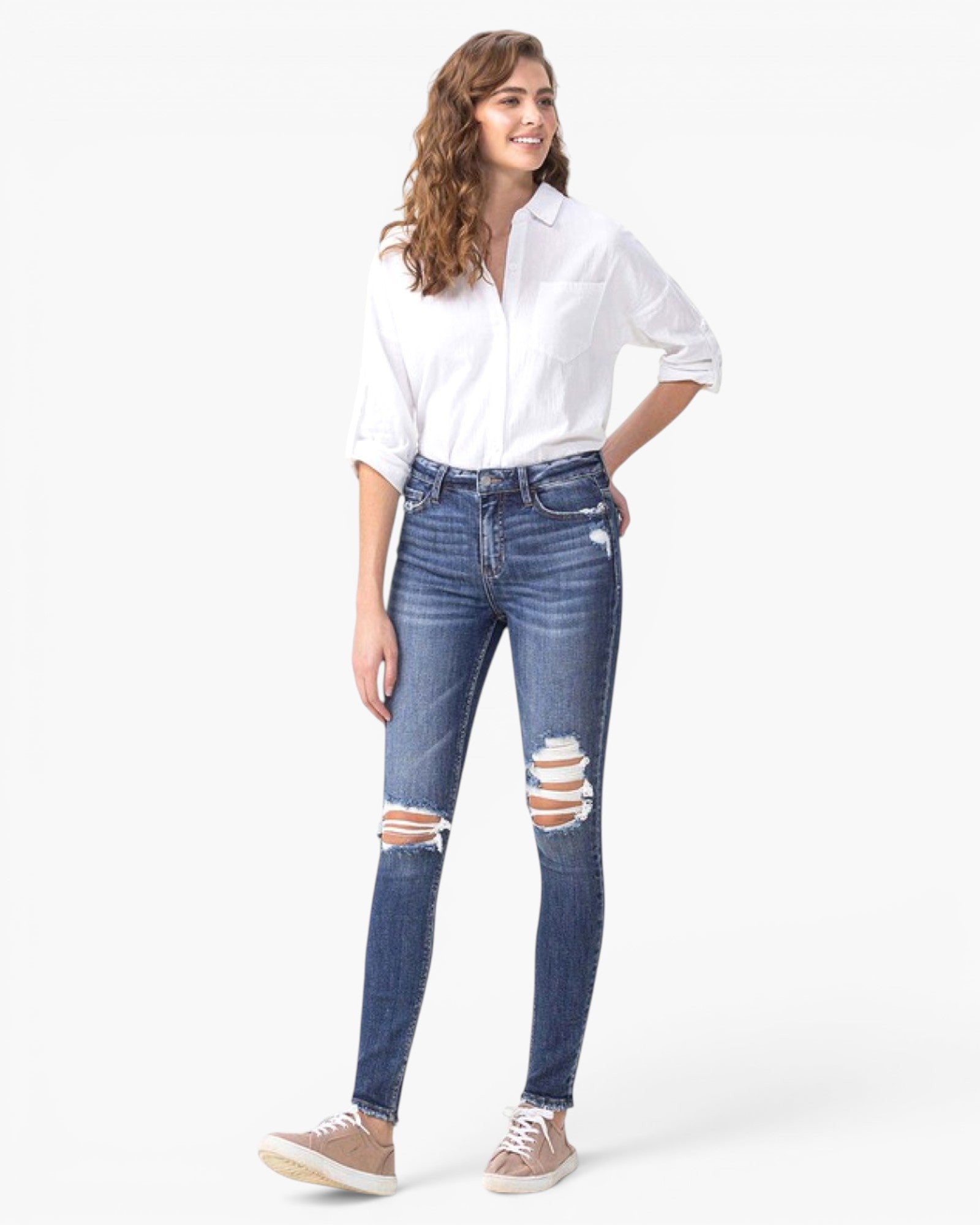A woman modeling a pair of high-waisted, blue skinny jeans with distressed rips at the knees, styled with a white button-down shirt and beige sneakers against a plain white background.