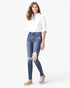 A woman modeling a pair of blue distressed skinny jeans with ripped knees, styled with a white button-down shirt and beige sneakers against a plain white background.