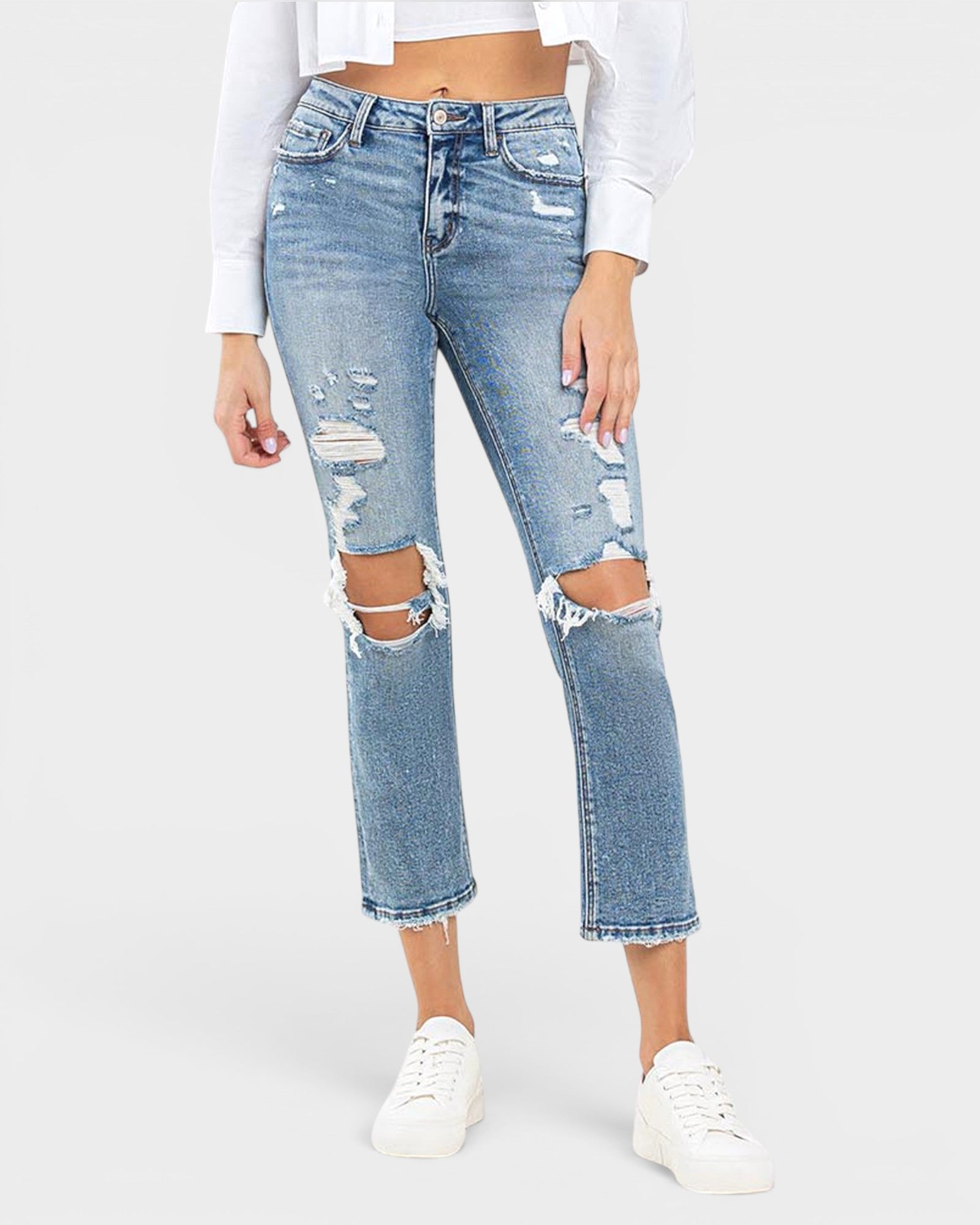 A model wearing light-wash blue distressed cropped jeans with ripped knees and frayed hems, paired with a white cropped top and white sneakers, against a light gray studio background.
