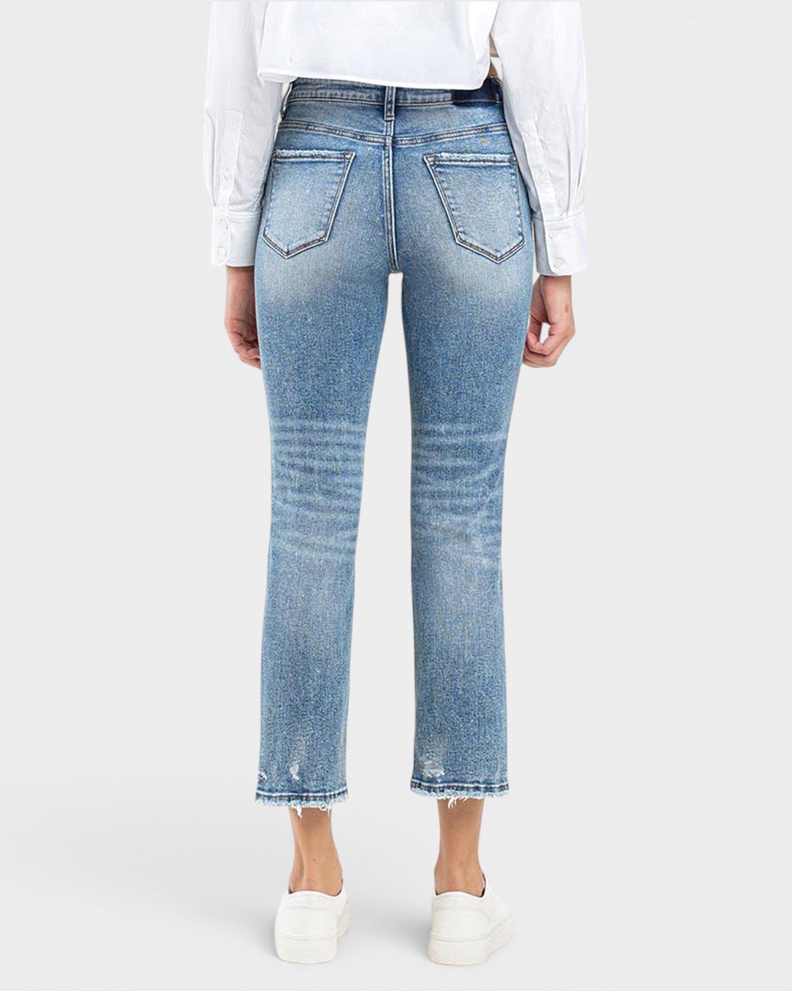 Rear view of a person wearing light-wash blue cropped jeans with a straight leg, frayed hems, and whiskering, paired with a white long-sleeve top and white slip-on shoes against a light gray studio background.