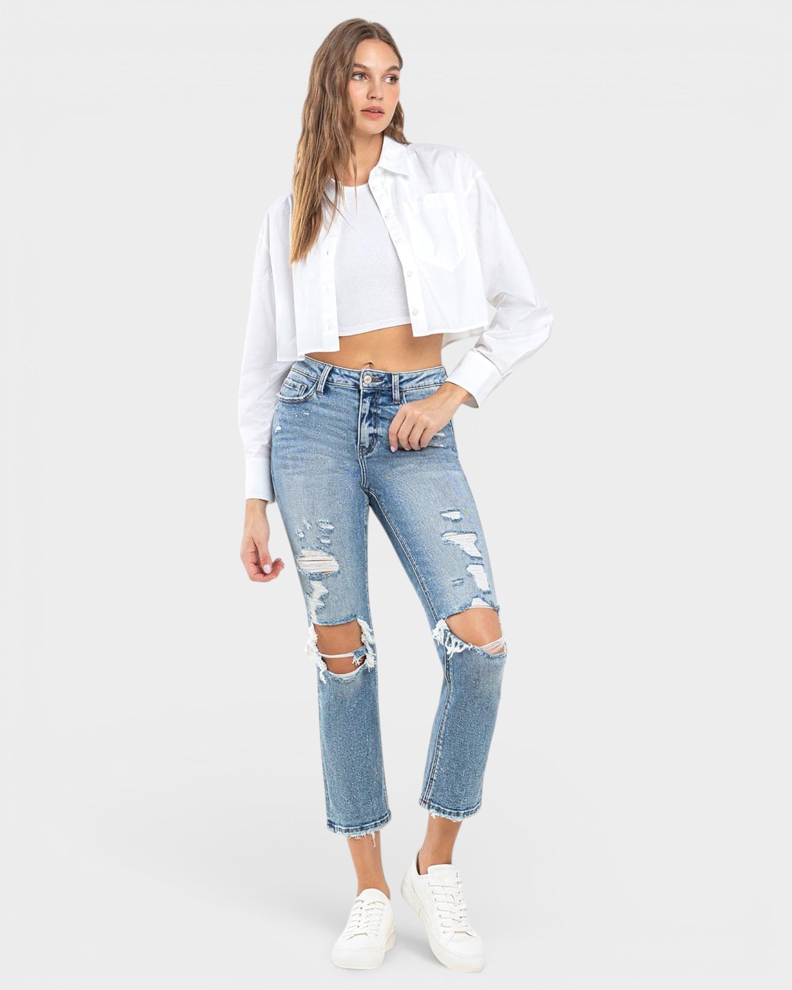 A model wearing light-wash blue distressed cropped jeans with ripped knees, styled with a white cropped tee, a white open-front shirt, and white sneakers against a light gray studio background.