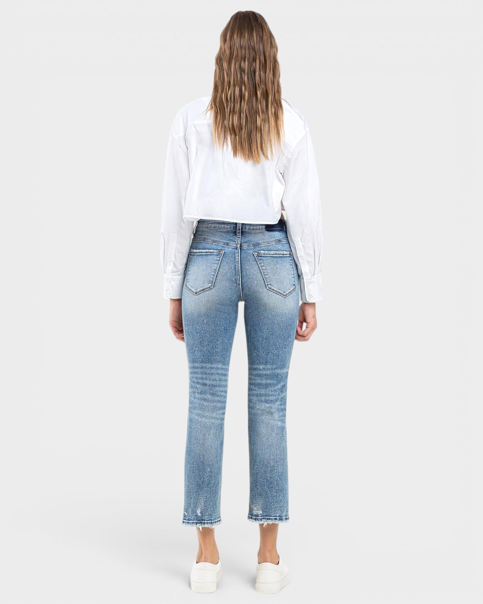A woman shown from the back wearing light-wash blue cropped jeans, a white long-sleeve shirt, and white sneakers against a light gray studio background.