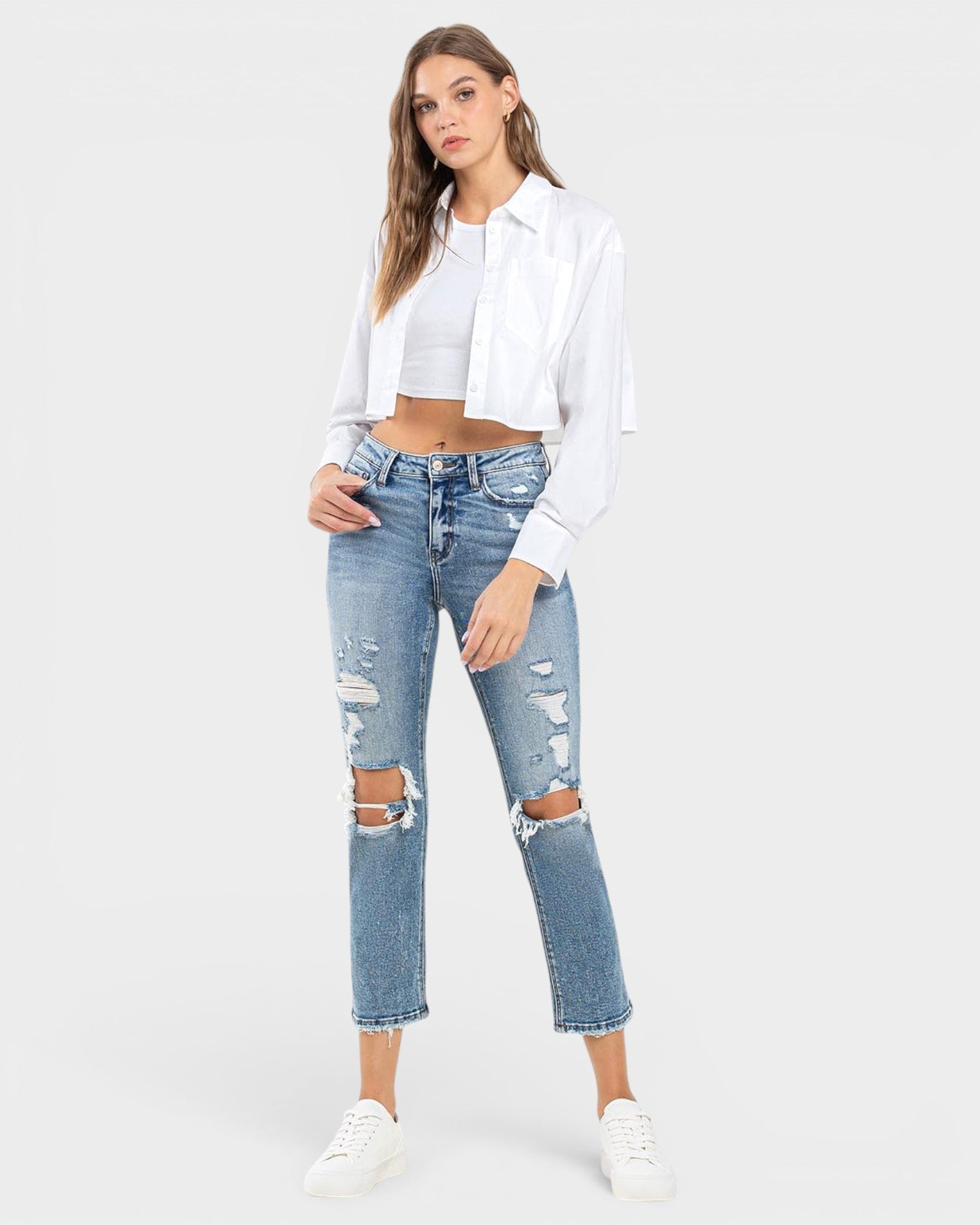 A model wearing light-wash blue distressed cropped jeans with ripped knee details, styled with a white cropped shirt, white crop top, and white sneakers against a light gray studio background.