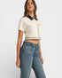 Woman wearing a white cropped top with black trim and blue jeans on a light gray background