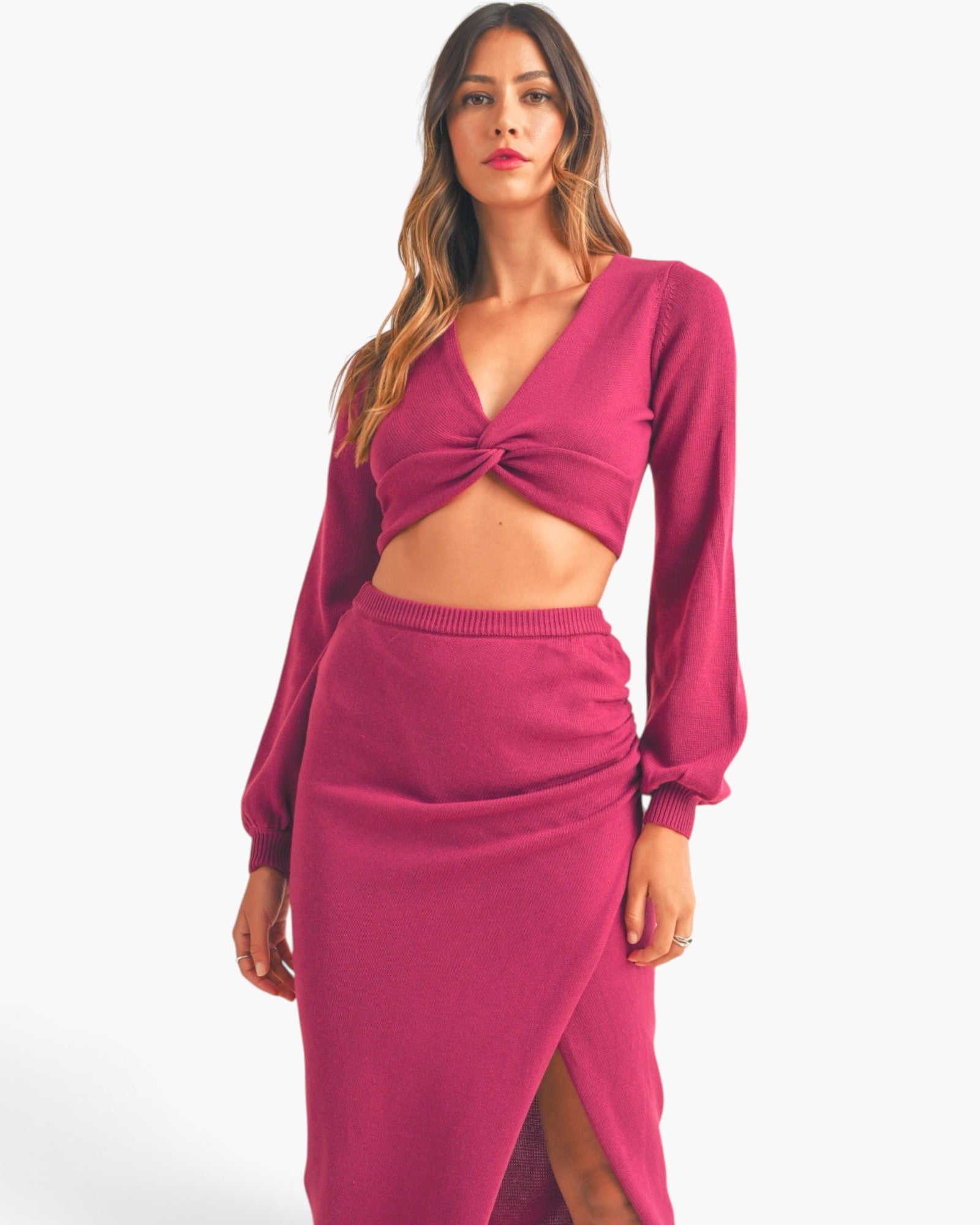 A woman modeling a matching knit two-piece set in a rich magenta pink, featuring a long-sleeve crop top with a twisted front detail and a high-waisted ruched skirt with a front slit, set against a plain light gray background.