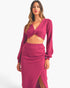 A woman modeling a matching knit two-piece set in a rich magenta pink, featuring a long-sleeve crop top with a twisted front detail and a high-waisted ruched skirt with a front slit, set against a plain light gray background.