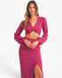 A woman modeling a matching two-piece knit set in a rich magenta pink, featuring a long-sleeve wrap-style crop top with a twisted front detail and a high-waisted wrap skirt with a thigh-high slit, set against a plain light gray studio background.