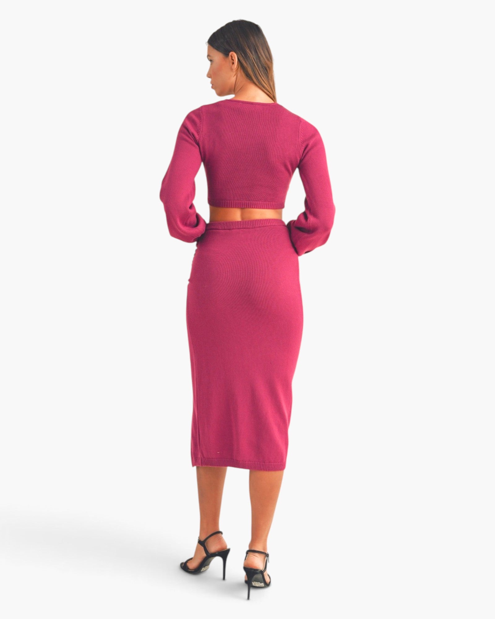 A woman shown from the back wearing a bright pink two-piece knit set consisting of a long-sleeve crop top and a matching midi skirt, paired with black high-heeled sandals against a light gray studio background.