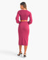 A woman shown from the back wearing a bright pink two-piece knit set consisting of a long-sleeve crop top and a matching midi skirt, paired with black high-heeled sandals against a light gray studio background.
