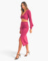 A woman modeling a bright pink two-piece knit set consisting of a long-sleeve wrap-style crop top and a matching high-waisted wrap skirt with a thigh slit, paired with black strappy high-heeled sandals against a light gray studio background.