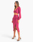 A woman modeling a bright pink two-piece outfit consisting of a long-sleeve wrap-style midi skirt and a matching twisted crop top, paired with black strappy high-heeled sandals against a light gray studio background.