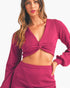A woman wearing a magenta knit long-sleeve crop top with a twisted front detail and matching knit bottoms, photographed against a plain light background.