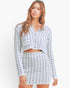 A woman modeling a matching two-piece knit set featuring a long-sleeve cropped button-front top and a high-waisted mini skirt, both in a light gray and white houndstooth pattern.