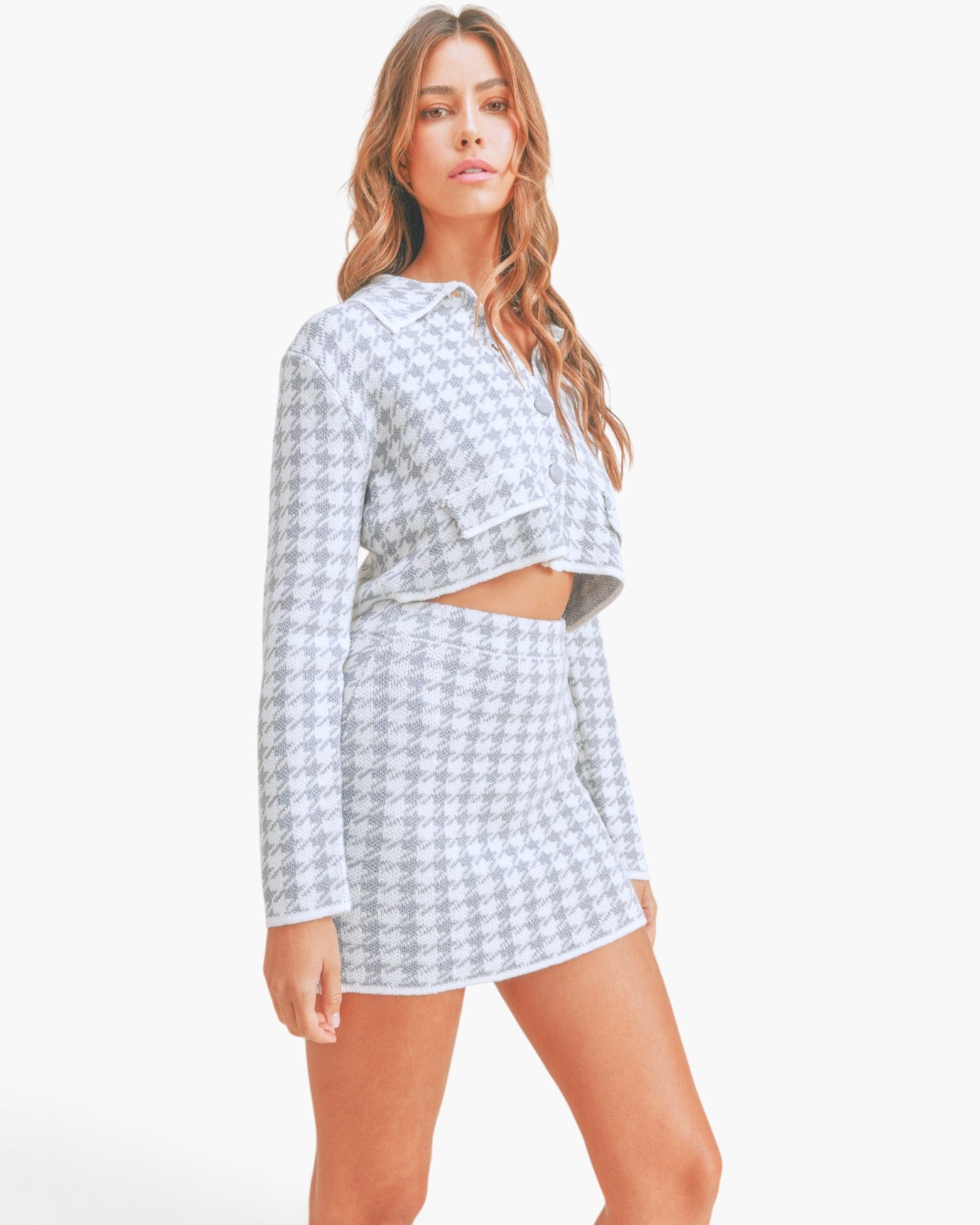 A woman modeling a matching two-piece outfit featuring a light gray and white houndstooth pattern, consisting of a long-sleeve cropped top and a high-waisted mini skirt, against a plain white studio background.