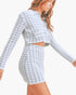 A woman wearing a matching long-sleeve collared crop top and high-waisted mini skirt set in a light gray and white houndstooth pattern, shown against a plain light background.