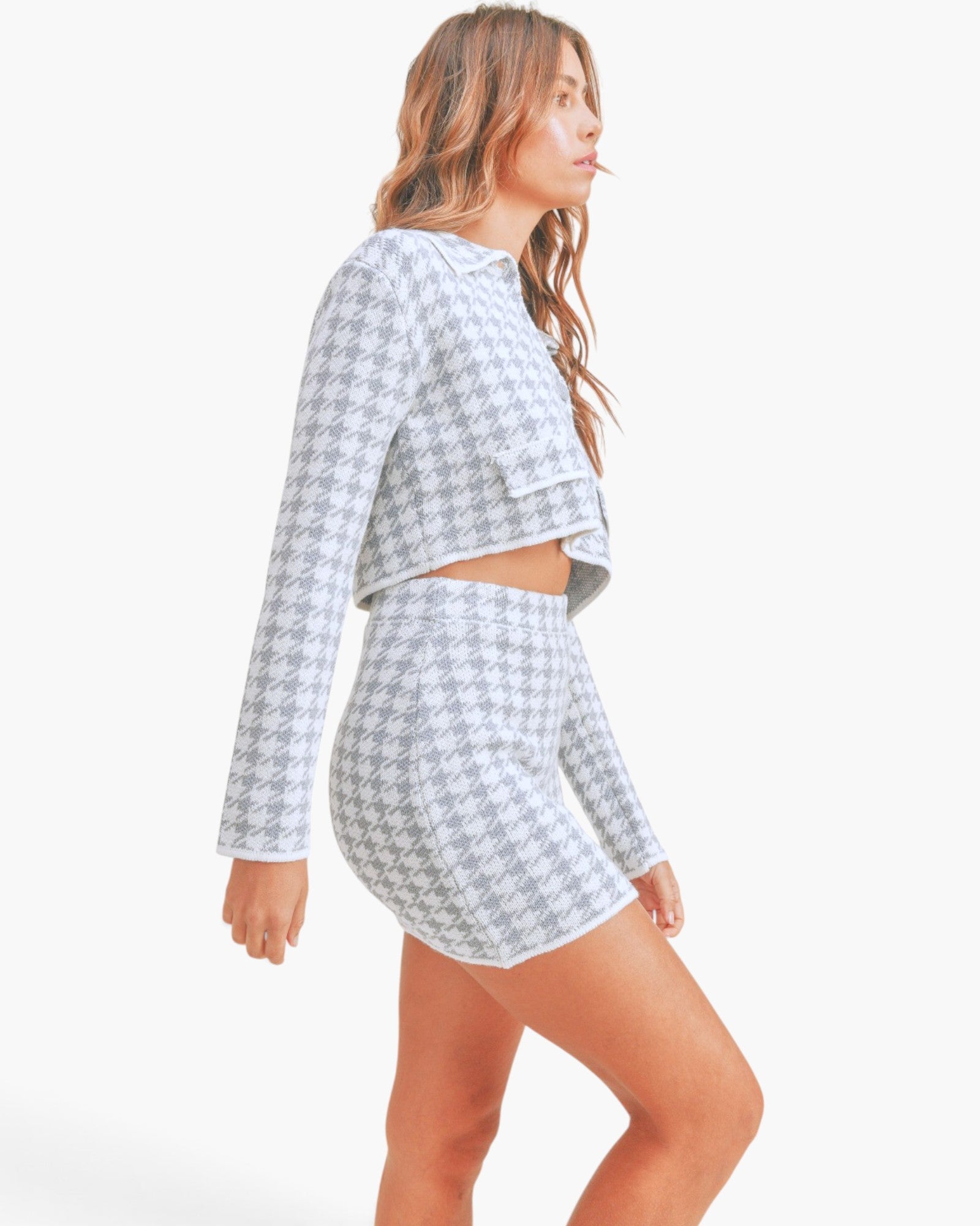 A woman modeling a matching two-piece knit set featuring a long-sleeve cropped top and high-waisted shorts in a light gray and white houndstooth pattern, set against a plain white background.