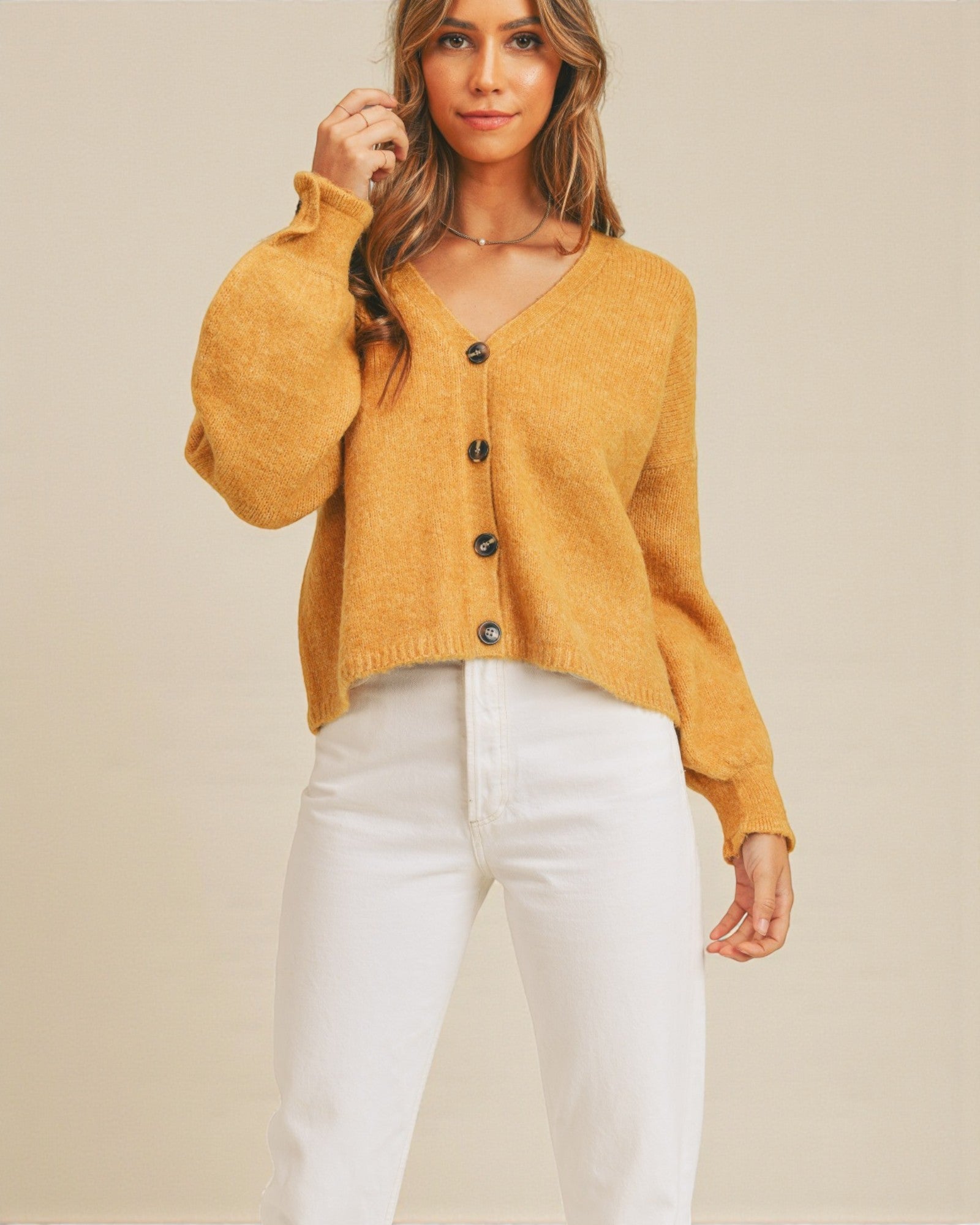 Woman wearing a mustard yellow cardigan with black buttons and white pants on a beige background
