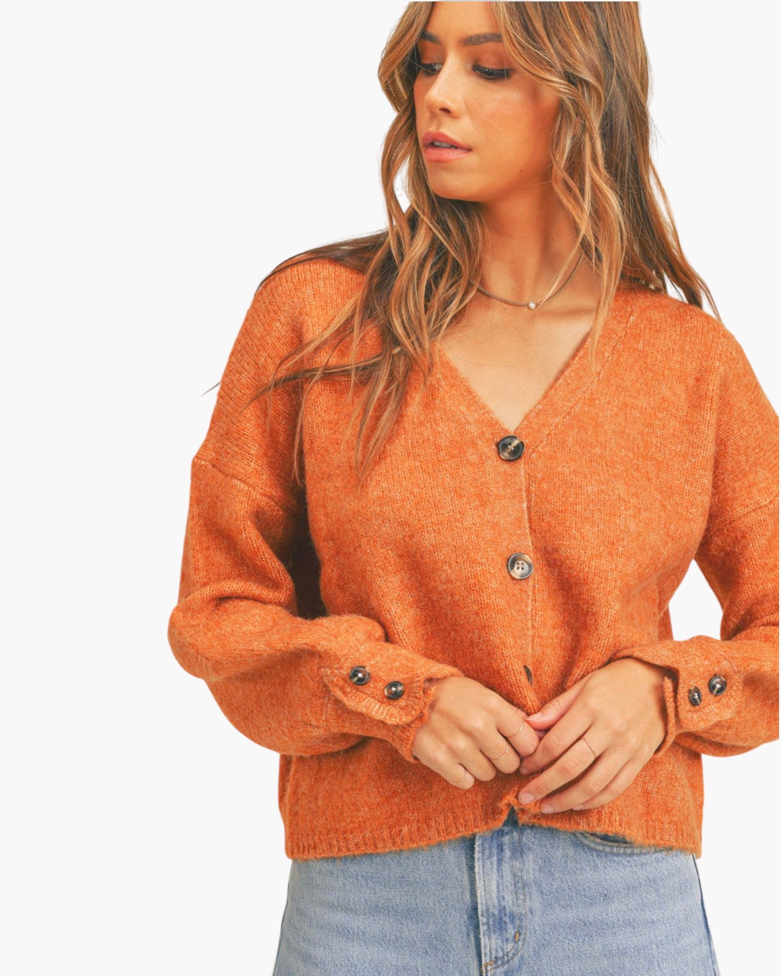 A woman wearing a fuzzy orange cardigan sweater with button details on the front and cuffs, paired with light blue jeans, against a plain white background.