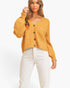 A woman modeling a cropped, mustard-yellow fuzzy knit cardigan with dark buttons, paired with white pants, against a clean white background.