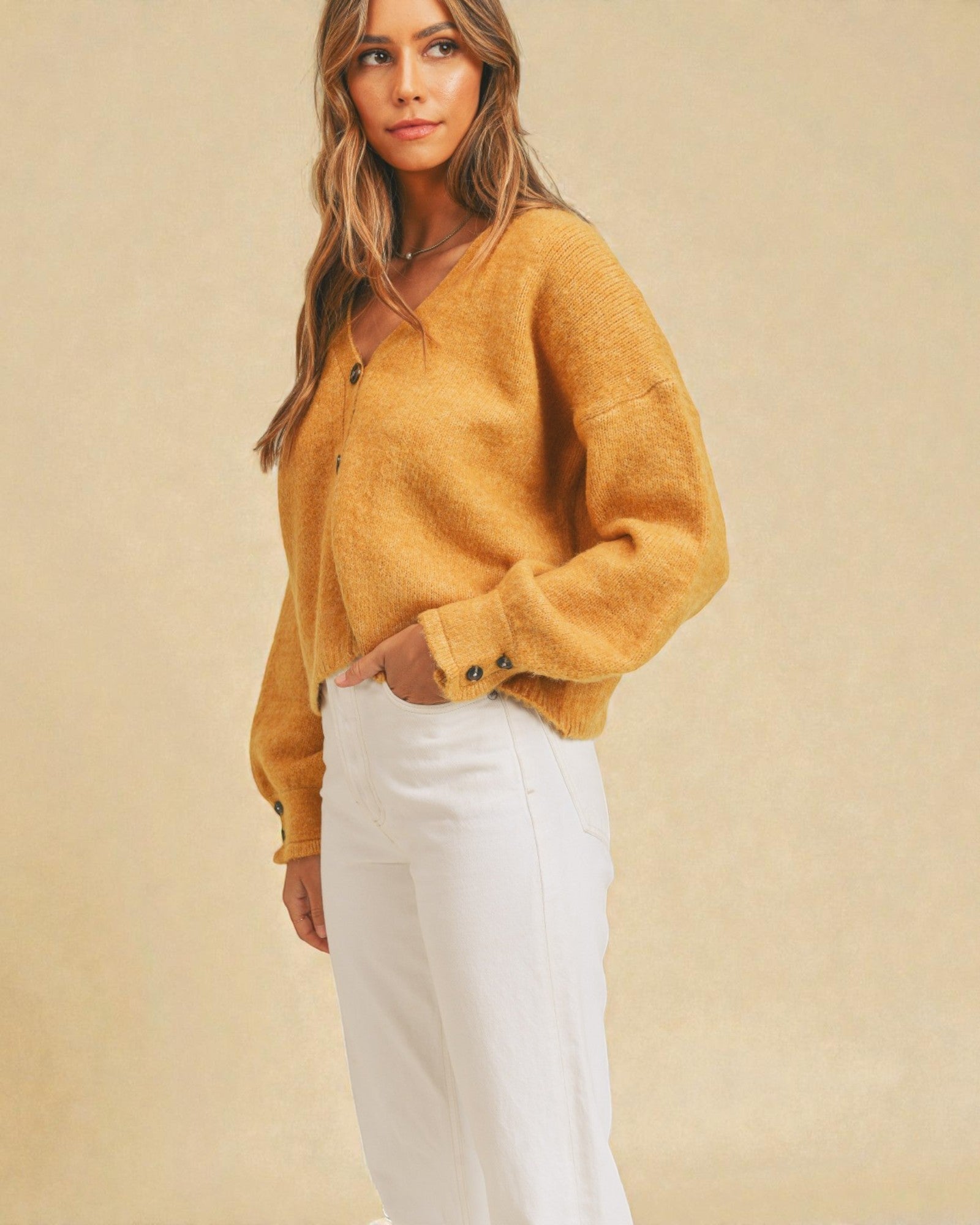 Woman wearing a mustard yellow cardigan and white pants against a beige background