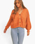 A woman wearing a fuzzy orange cropped cardigan with a V-neck and button-front closure, paired with light blue jeans, against a plain white background.
