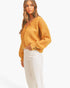 A woman wearing a fuzzy mustard yellow button-up cardigan sweater and white pants, photographed against a clean white studio background.