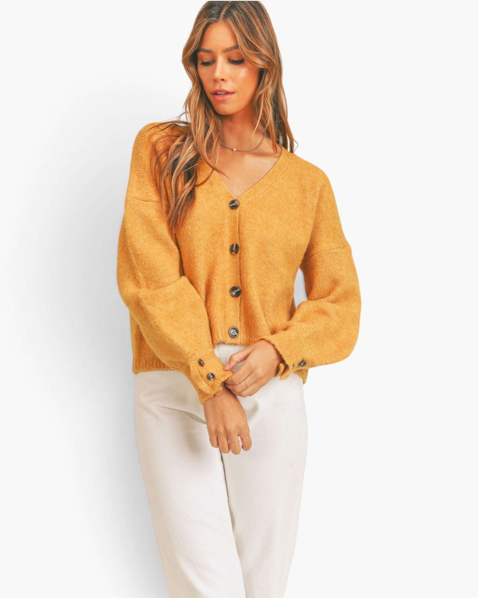 A woman wearing a mustard yellow fuzzy knit cardigan with dark buttons and white pants, standing against a plain white studio background.