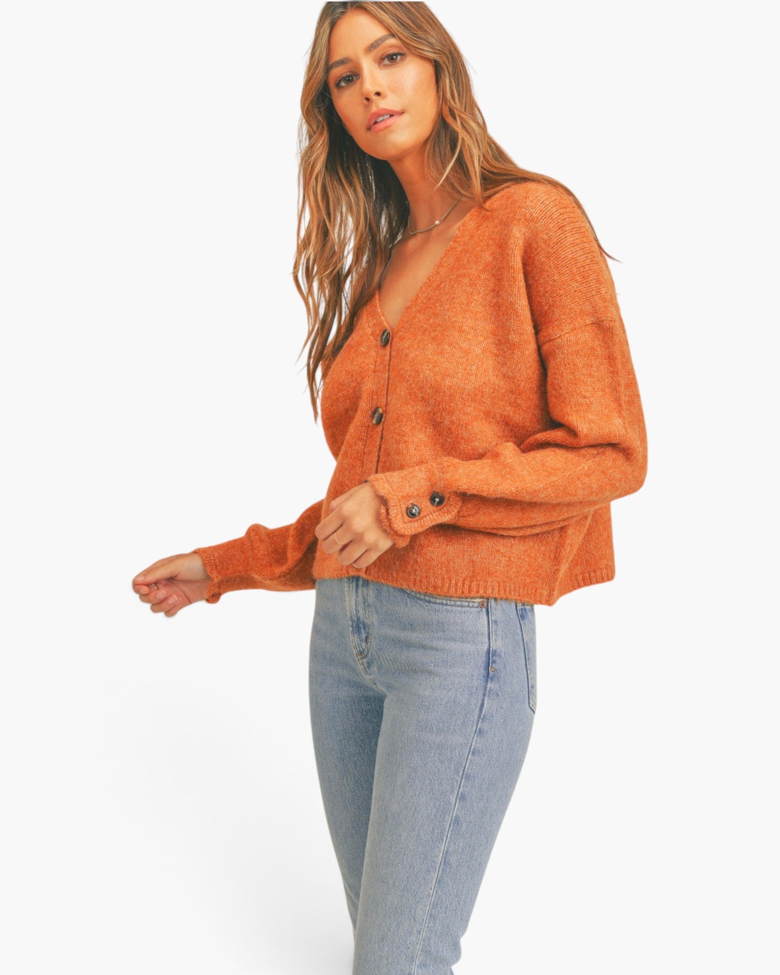 A woman modeling a cropped, long-sleeve knit cardigan in a warm burnt orange color, paired with light-wash blue jeans against a plain white studio background.