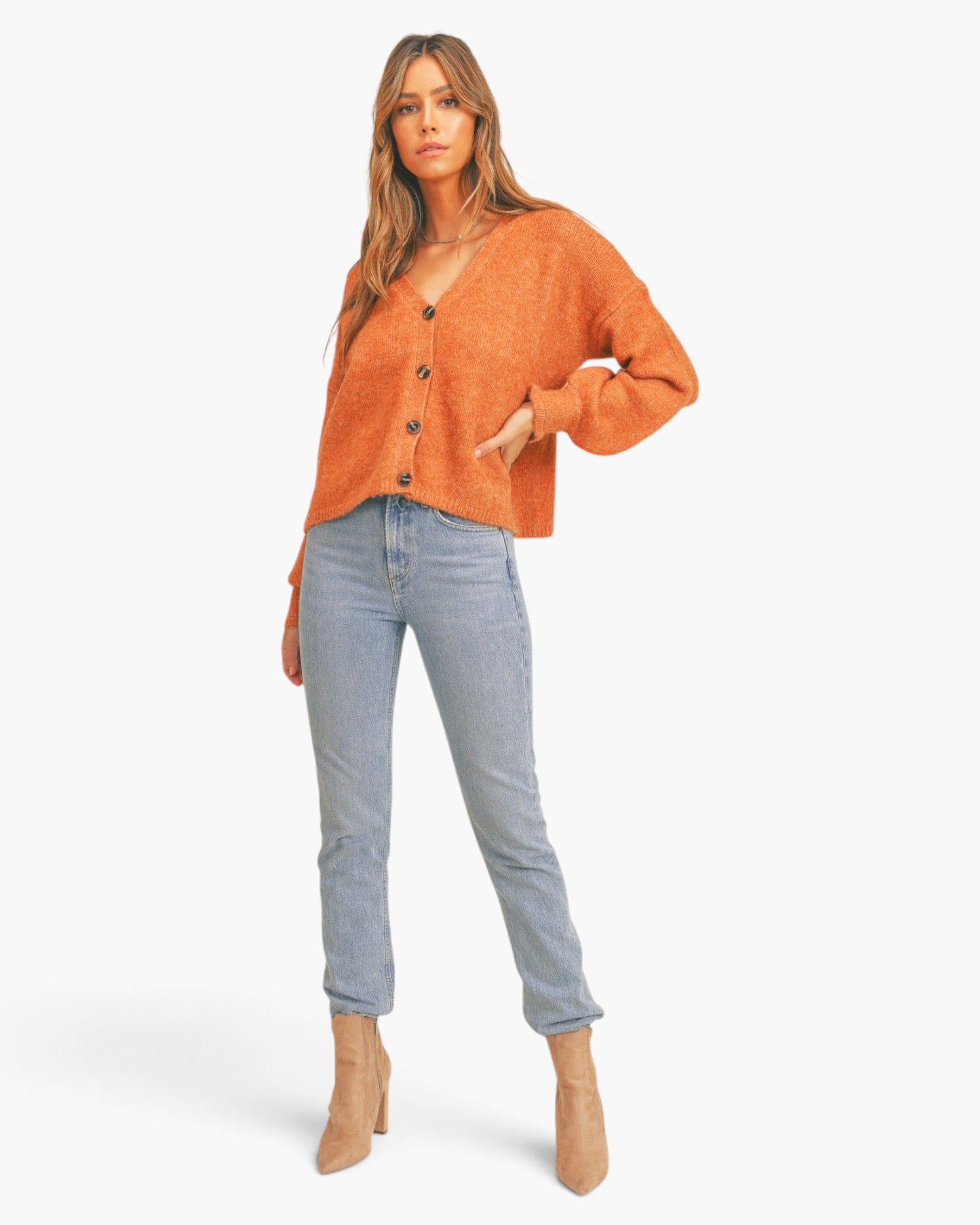 A woman modeling a cropped, long-sleeve orange knit cardigan with dark buttons, paired with light-wash blue jeans and beige ankle boots, against a plain white studio background.