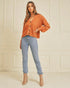 Woman wearing a rust-colored sweater and light blue jeans against a beige wall.