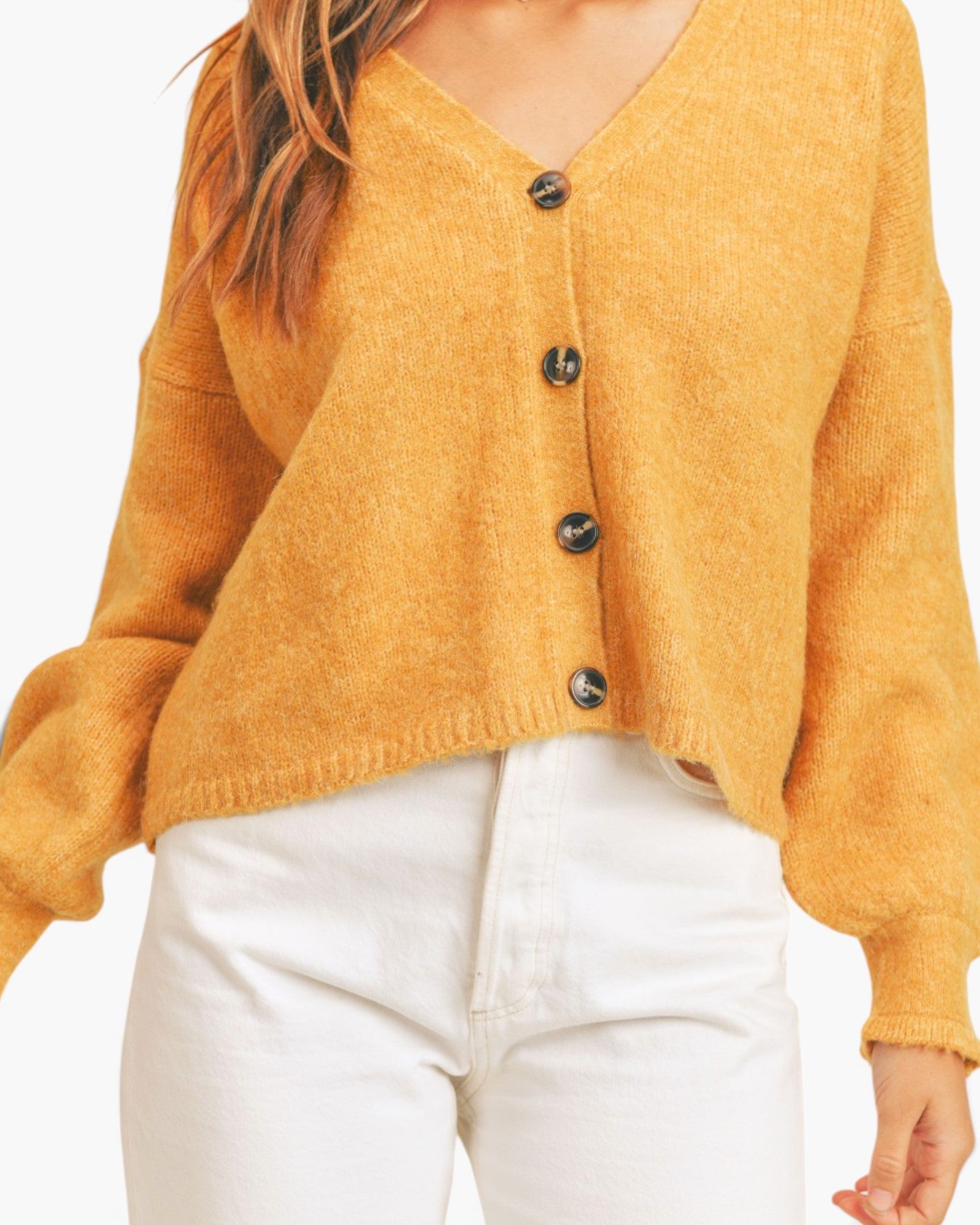 A person wearing a fuzzy mustard yellow cardigan with dark buttons and white pants, shown from the shoulders to the hips against a plain light background.