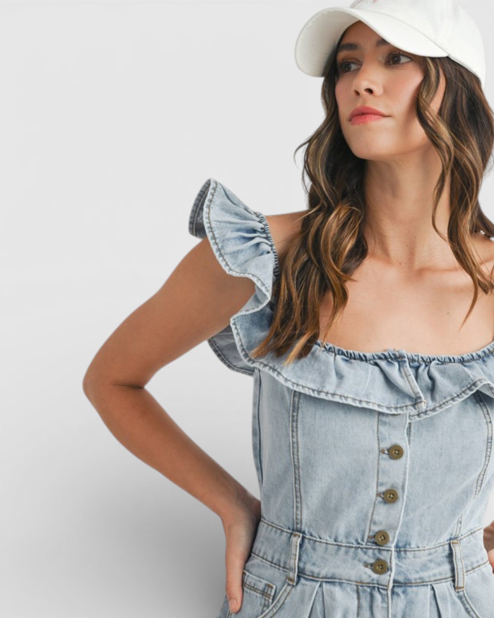 Woman wearing a light blue denim dress with ruffled details and a white cap on a plain background