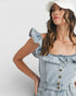 Woman wearing a light blue denim dress with ruffled details and a white cap on a plain background