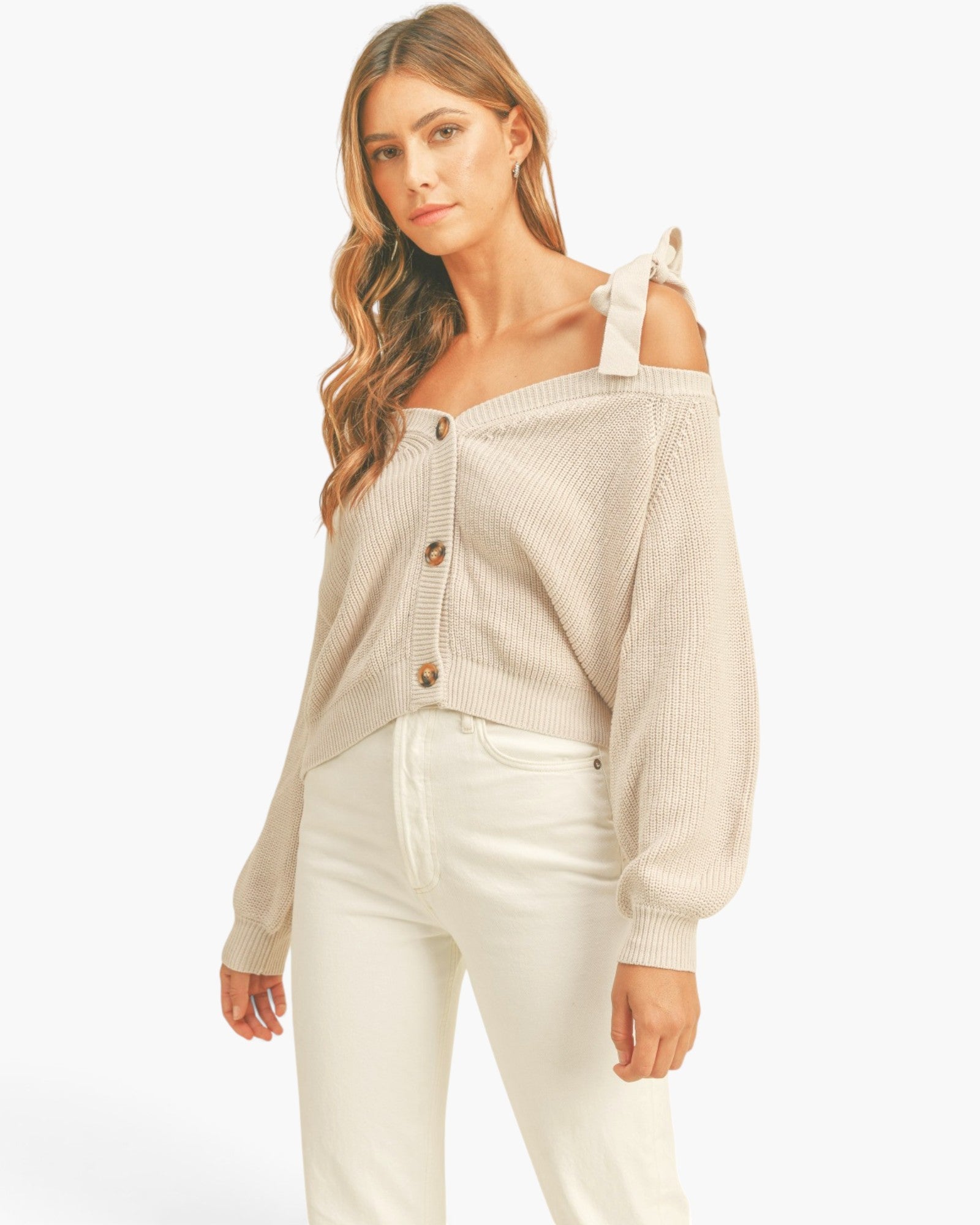 A woman modeling a beige ribbed knit cardigan with tie-shoulder straps and brown buttons, paired with white pants, against a plain white studio background.