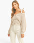 A woman modeling a beige ribbed knit cardigan with tie-shoulder straps and brown buttons, paired with white pants, against a plain white studio background.