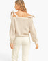 A woman shown from the back wearing a ribbed knit off-the-shoulder sweater with tie straps and white pants, against a plain light gray studio background.
