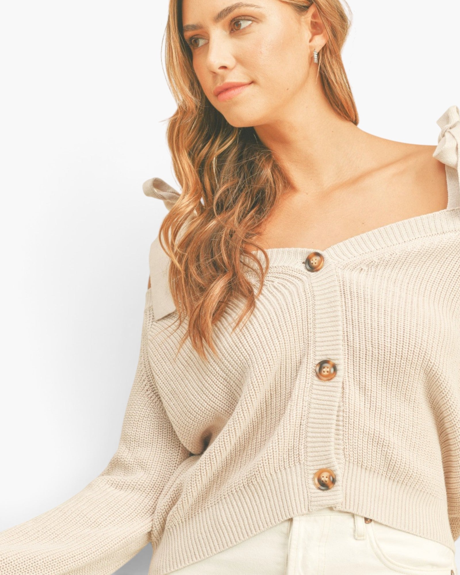 A woman wearing a ribbed knit off-the-shoulder cardigan in a light beige/cream color with brown buttons, paired with white pants against a plain white background.