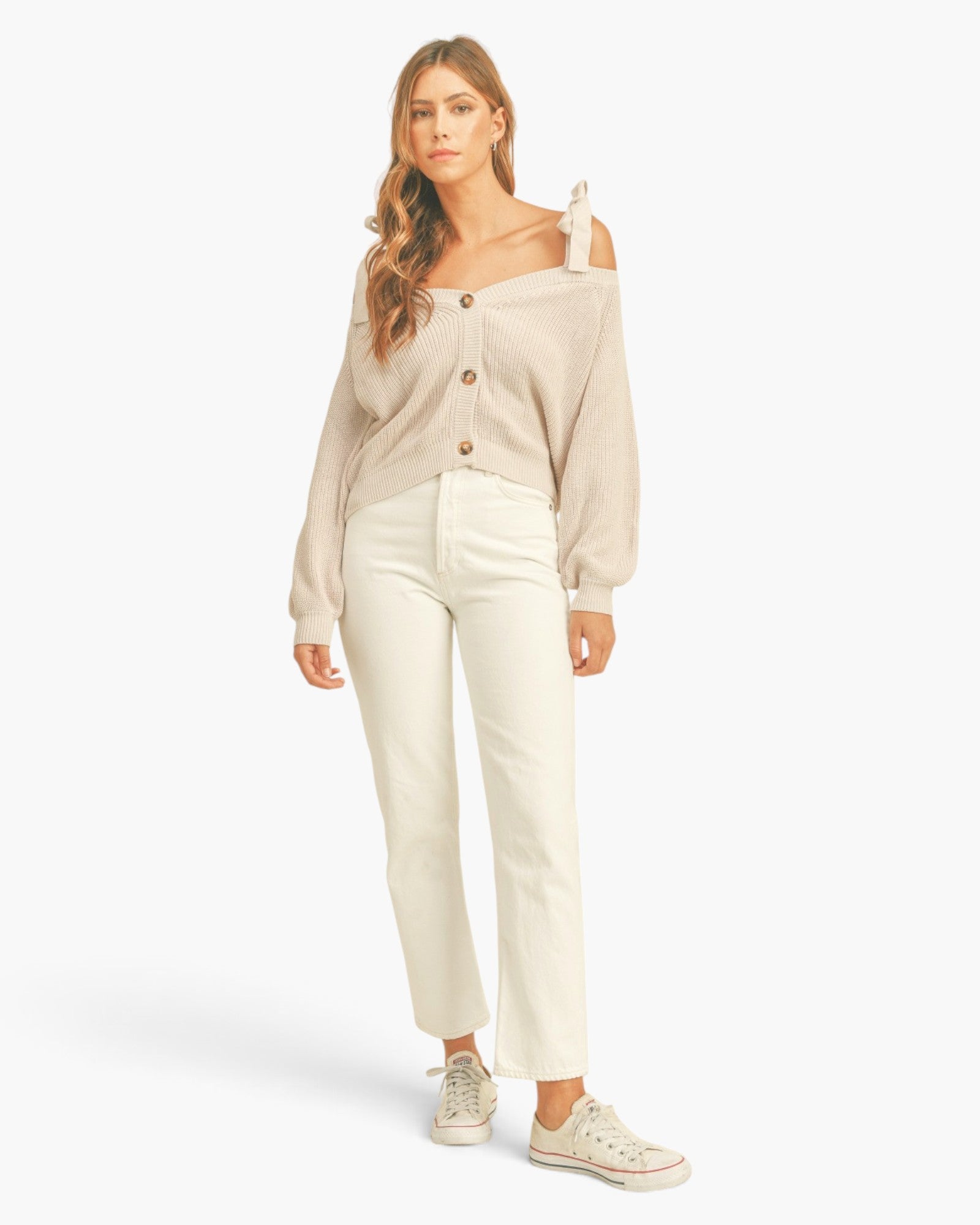 A woman modeling a beige off-the-shoulder button-front cardigan with cream-colored pants and light-colored sneakers, set against a plain white studio background.