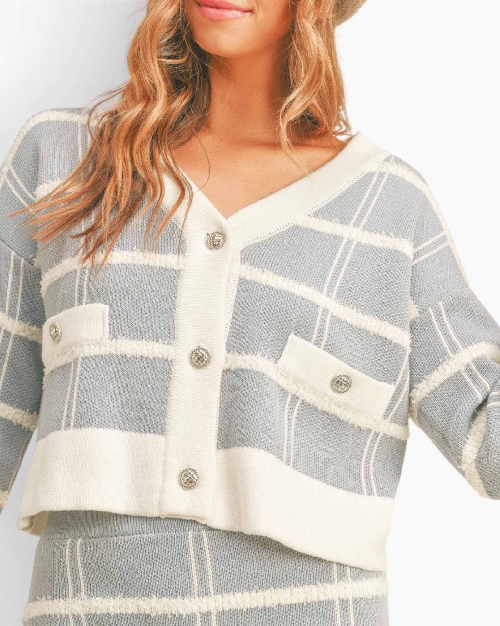 A close-up of a woman wearing a cropped knit cardigan with a gray and off-white plaid pattern, featuring a frayed trim and decorative buttons.