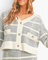 A close-up of a woman wearing a cropped knit cardigan with a gray and off-white plaid pattern, featuring a frayed trim and decorative buttons.