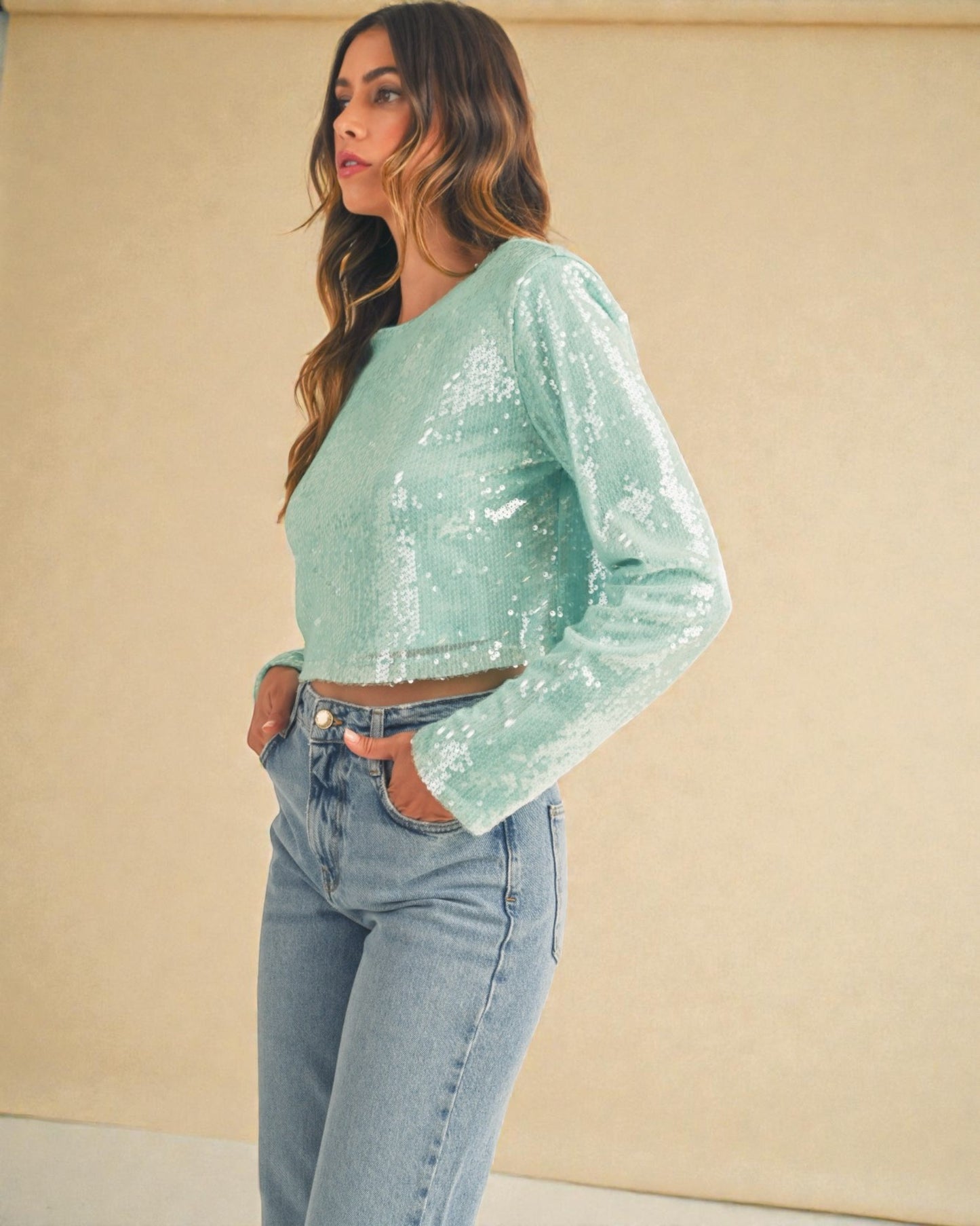 Woman wearing a light green sequin top and blue jeans against a beige background