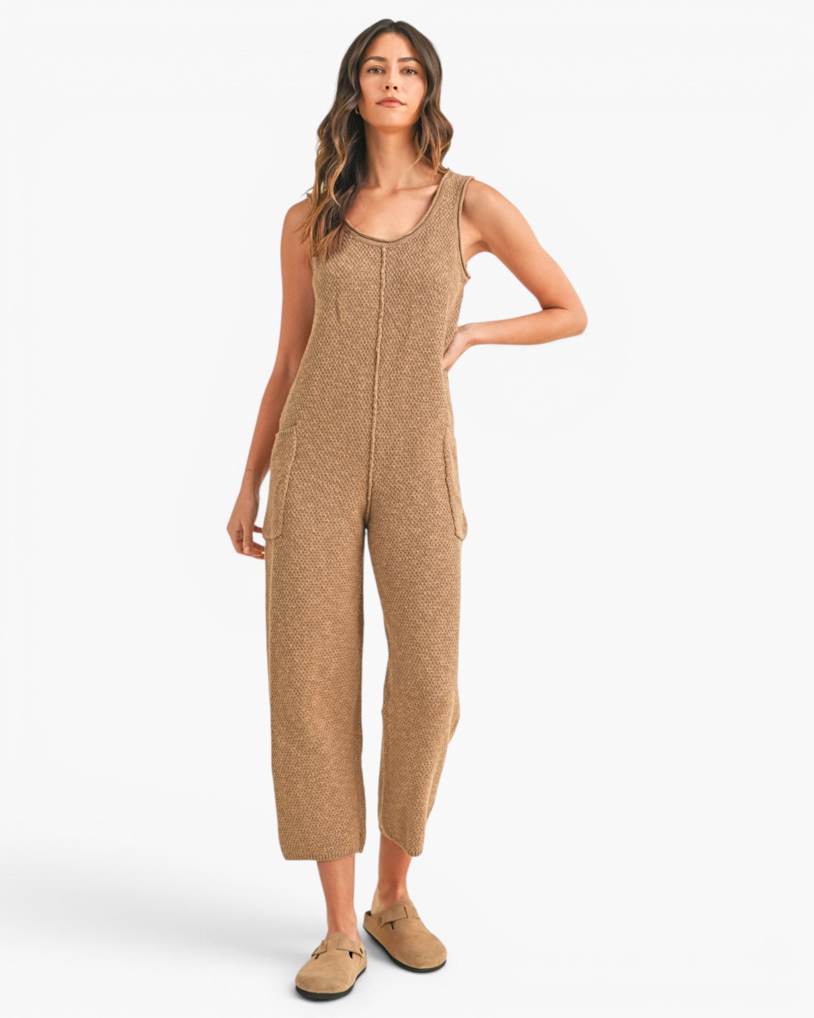 A woman modeling a sleeveless, cropped knit jumpsuit in a warm tan color, paired with matching tan slip-on shoes, against a plain white studio background.