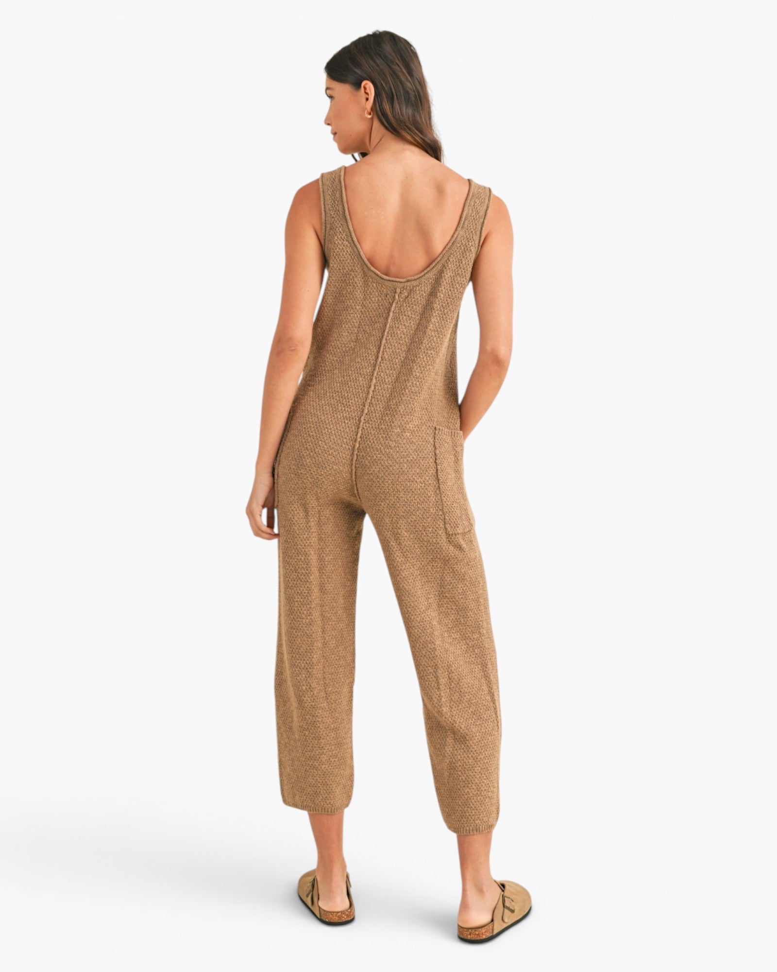 A woman shown from the back wearing a sleeveless, cropped jumpsuit in a heathered tan-brown color, paired with flat tan sandals, against a plain white studio background.