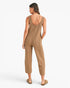 A woman shown from the back wearing a sleeveless, cropped jumpsuit in a heathered tan-brown color, paired with flat tan sandals, against a plain white studio background.