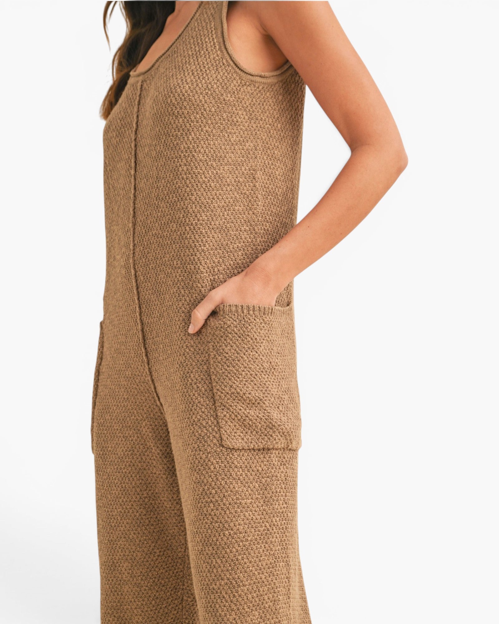 A close-up side view of a woman wearing a textured knit sleeveless jumpsuit in a warm camel brown color, with a side pocket and a visible front zipper.