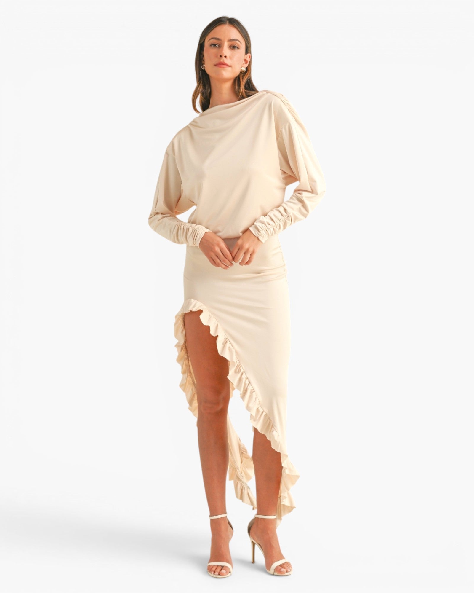 A model wearing a beige draped long-sleeve midi dress with a ruffled high-low hem and gathered sleeves, paired with white strappy heeled sandals, against a plain white studio background.