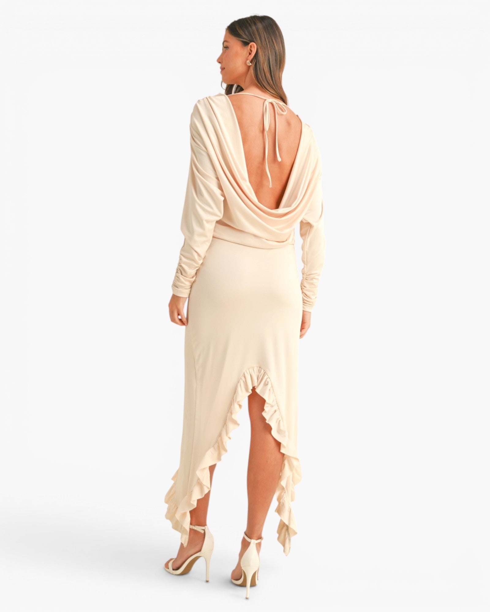 A woman shown from the back wearing a long-sleeve cream maxi dress with a draped open back, ruffled hem, and high-low front slit, paired with nude high heels against a plain white studio background.