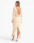 A woman shown from the back wearing a long-sleeve cream maxi dress with a draped open back, ruffled hem, and high-low front slit, paired with nude high heels against a plain white studio background.