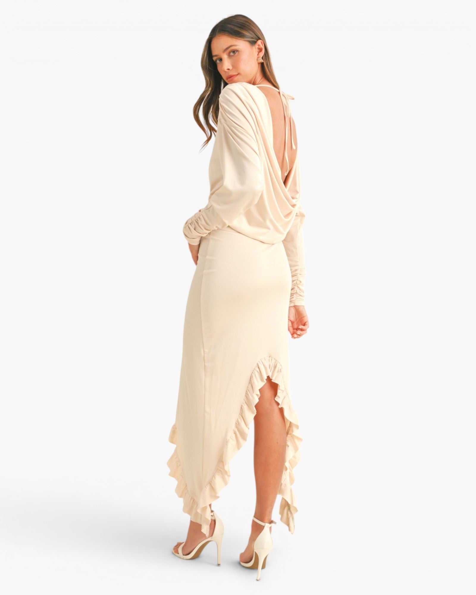 A woman modeling a long-sleeve cream maxi dress with a draped open back and ruffled hem, shown from the back against a plain white studio background.